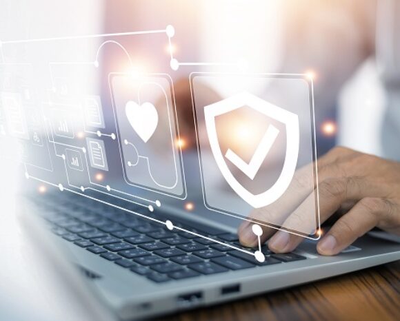 Eine Person tippt auf einem Laptop. Über der Tastatur befinden sich holografische Symbole, darunter ein Schild mit einem Häkchen. Die Szene vermittelt ein digitales Sicherheitsthema und spiegelt die Ergebnisse der Studie „Veraltete IT-Systeme bedrohen die Zukunft des Gesundheitswesens“ wider. Dabei stehen Sicherheit und Schutz im Vordergrund.