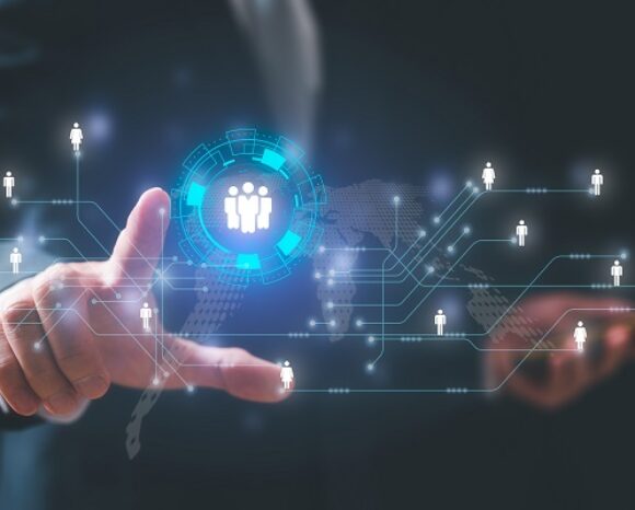 Eine Person im Anzug zeigt auf eine digitale Schnittstelle, auf der ein leuchtendes Symbol einer Gruppe von Menschen angezeigt wird, die von miteinander verbundenen Figuren umgeben sind. Dies symbolisiert vor einem dunklen Hintergrund „BSI goes USA: Projektarbeit in San Francisco gestartet“ in den sozialen Netzwerken.