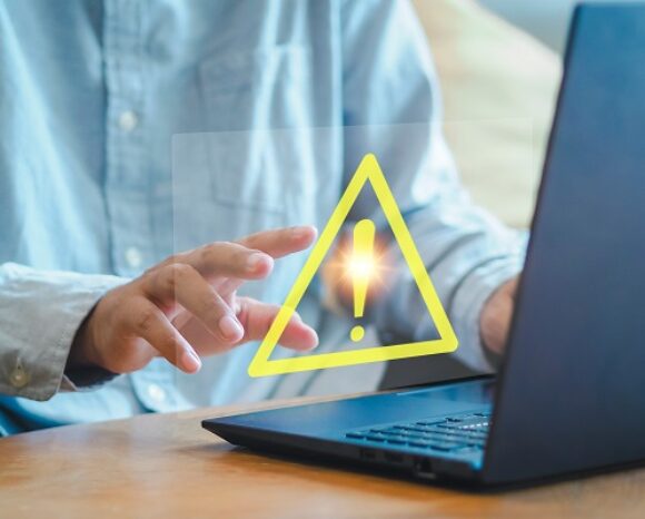 Mitarbeiter tippt auf Laptop-Tastator. Gelbes Dreieck mit Ausrufezeichen im Vordergrund