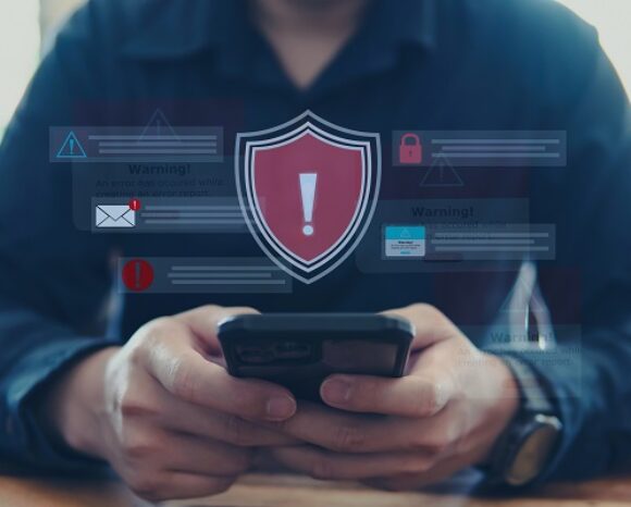 Eine Person hält ein Smartphone, über dem verschiedene digitale Warnsymbole angezeigt werden, darunter ein rotes Schild mit einem Ausrufezeichen. Die Person trägt ein dunkles Hemd. Der Fokus liegt auf dem Telefon und den Warnsymbolen.