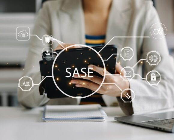 Eine Person im Blazer hält ein Tablet mit holografischen Symbolen wie einer Wolke, einem Schloss und WLAN. Der zentrale Text lautet „SASE“. Auf dem Schreibtisch davor stehen ein Laptop und ein Notebook.