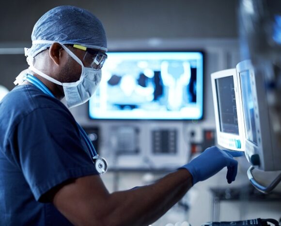Ein medizinischer Mitarbeiter in OP-Kleidung, Maske und Haube bedient medizinische Geräte in einem Krankenhaus. Mit Handschuhen und Stethoskop blickt er auf einen Monitor, der nahtlos integrierte medizinische Daten aus der Praxis in die Cloud anzeigt. Ein unscharfer Hintergrund suggeriert eine klinische Umgebung.