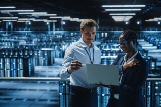 Frau und Mann (IT-Spezialisten) sprechen in einem Serverraum miteinander mit einem Laptop in der Hand