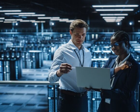 Frau und Mann (IT-Spezialisten) sprechen in einem Serverraum miteinander mit einem Laptop in der Hand
