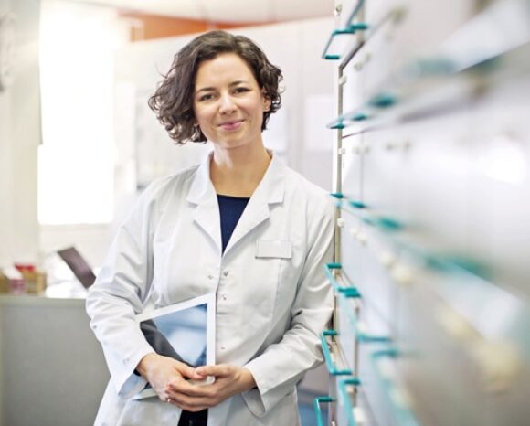 Junge Frau aus dem medizinischen Bereich lehnt an einer Tafel mit medizinischem Background und mit Tablet unter dem Arm